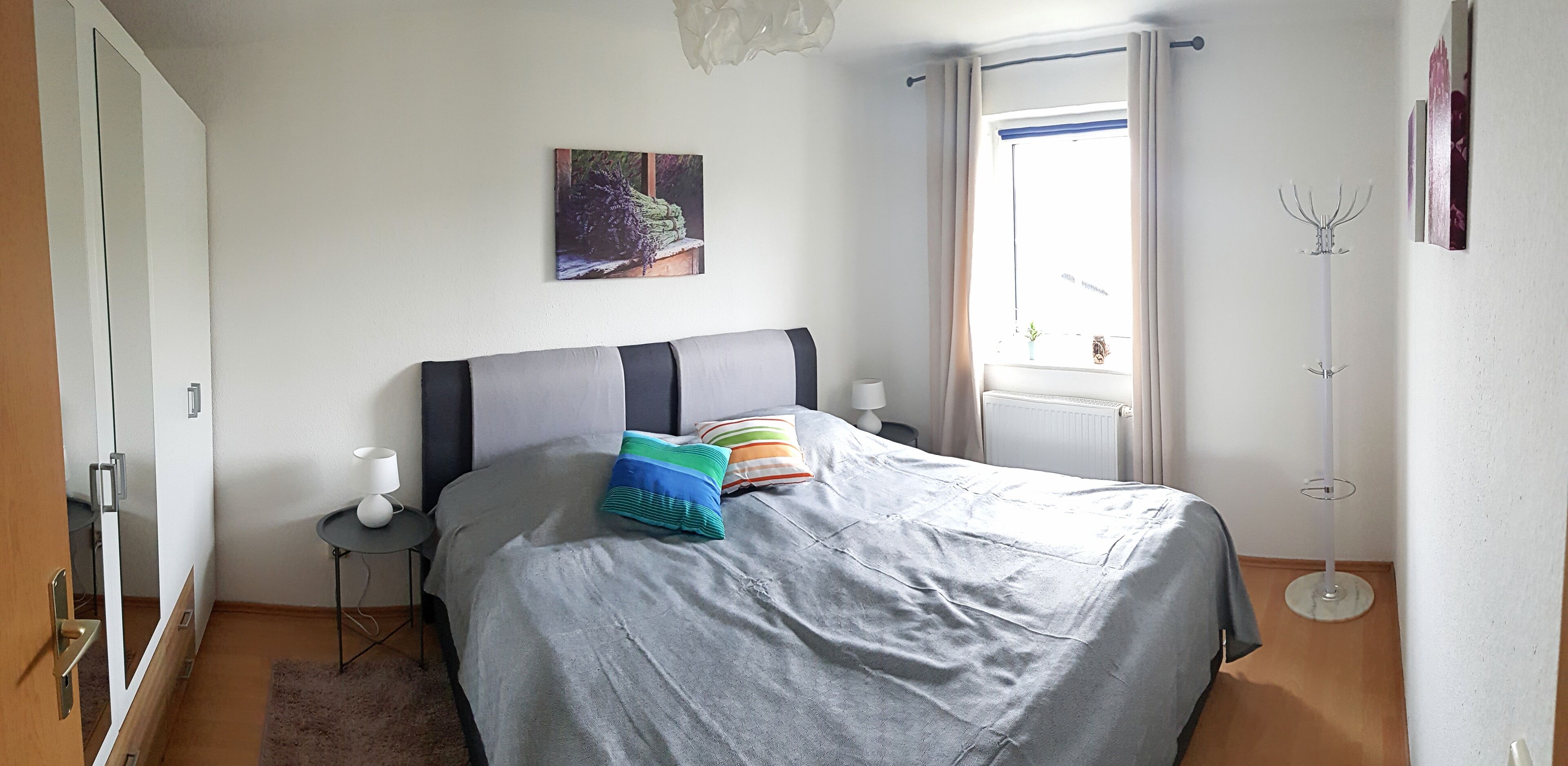 Ein Schlafzimmer mit Bett, Nachttisch, Lampe, einem Bild an der Wand und einem Fenster mit Vorhängen.