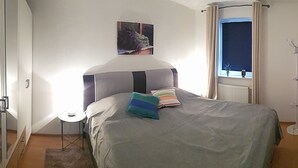 A bedroom with a bed, a small bedside table, a window with curtains, a radiator, and a wall-mounted light fixture. - Holidays directly in the spa town of Oberwiesenthal in a modern, cozy 3-room apartment (Kurort Oberwiesenthal)