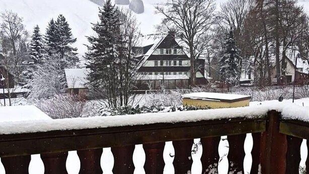A snow-covered balcony with a wooden railing, a view of a building with a sloped roof, and snow-laden trees in the background. - Holidays directly in the spa town of Oberwiesenthal in a modern, cozy 3-room apartment (Kurort Oberwiesenthal)