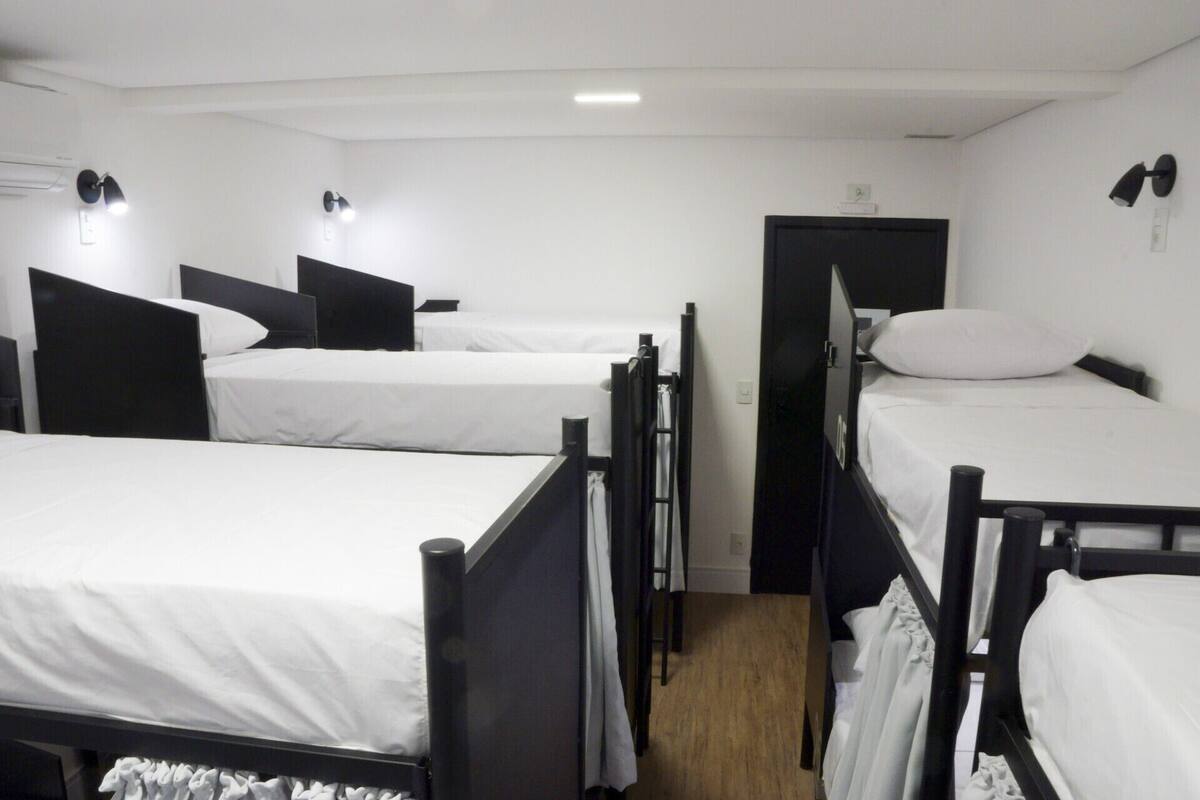 A room with four single beds arranged in a row, each with a black headboard and white bedding. There are wall-mounted lights above each bed.