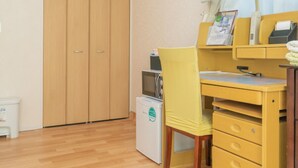 Una habitación compacta con un armario de madera, un escritorio amarillo con cajones, un refrigerador blanco pequeño y una ventana con cortinas.