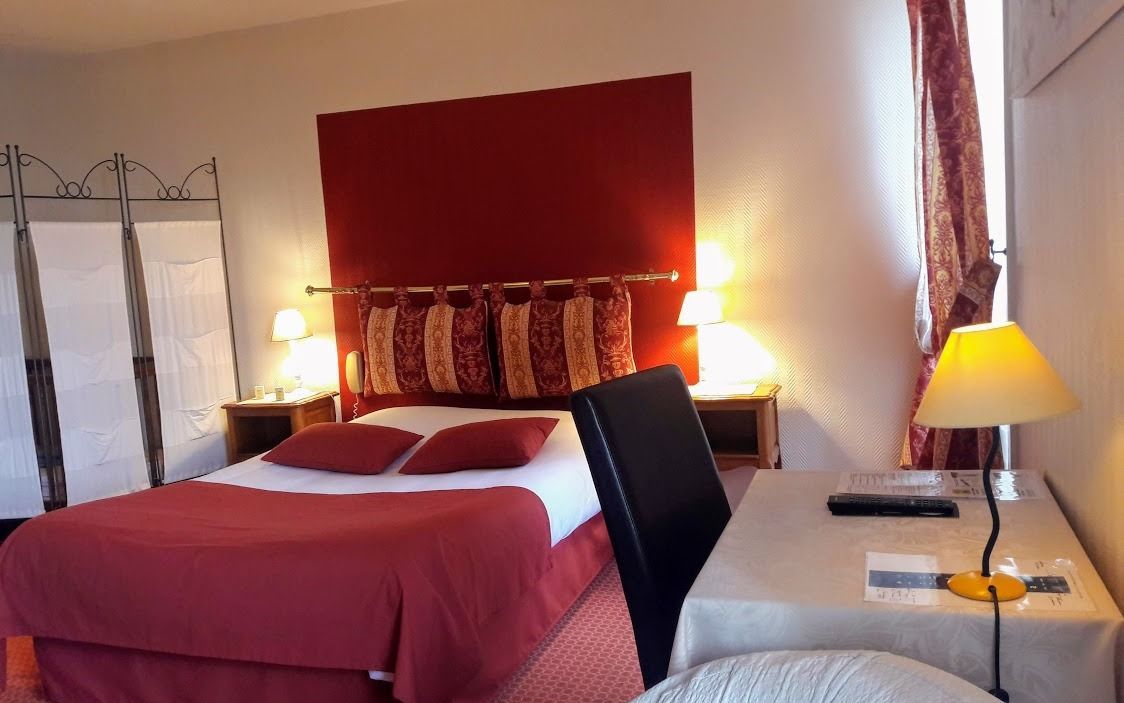 A hotel room with a bed, two bedside tables, a desk with a chair, and a red headboard.