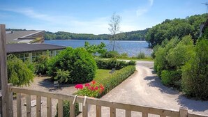 Vista de una propiedad junto al lago con camino de grava que conduce a una terraza de madera, rodeada de abundante vegetación y un cielo azul despejado.