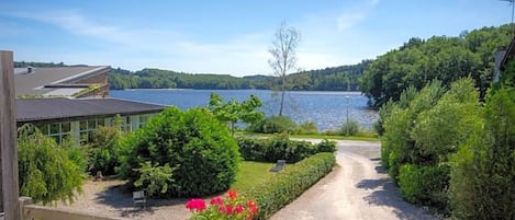 Vista de una propiedad junto al lago con camino de grava que conduce a una terraza de madera, rodeada de abundante vegetación y un cielo azul despejado.