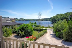 A view of a lakeside property with a gravel driveway leading to a wooden deck, surrounded by lush greenery and a clear blue sky.