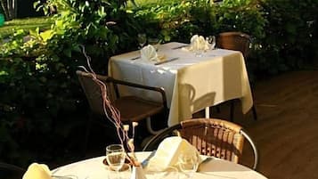 An outdoor dining area with round tables set for two, covered with white tablecloths, and surrounded by lush greenery.