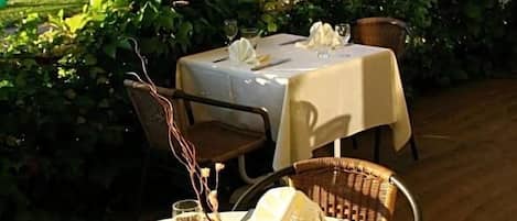 Un área de comedor al aire libre con mesas redondas dispuestas para dos, cubiertas con manteles blancos y rodeadas de abundante vegetación.