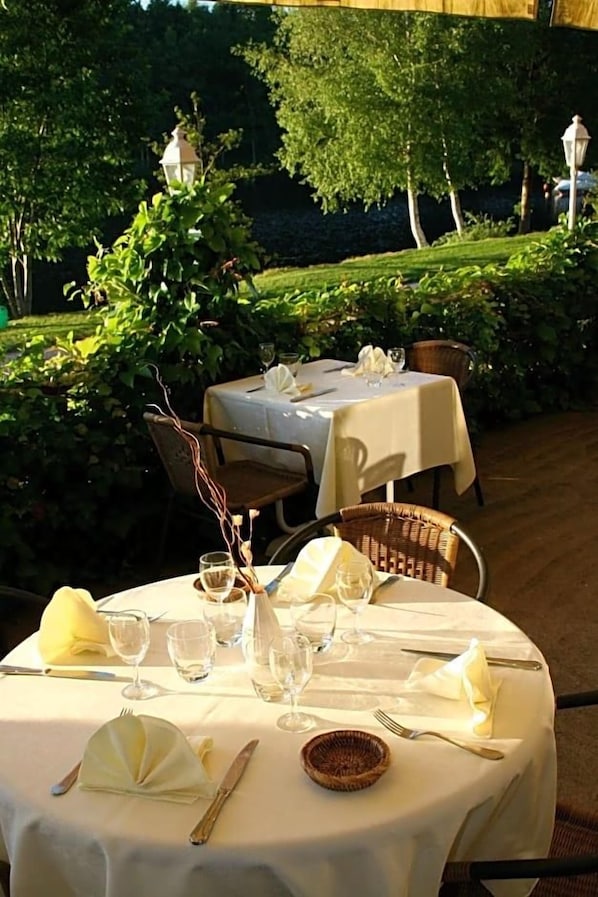 An outdoor dining area with round tables set for two, covered with white tablecloths, and surrounded by lush greenery. - Hotel du Lac (Neuvic)