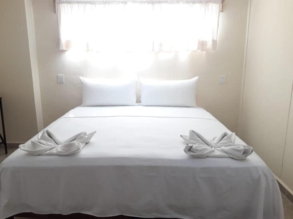 Una cama bien hecha con sábanas y almohadas blancas, una ventana con cortinas traslúcidas y un diseño de pared sencillo.