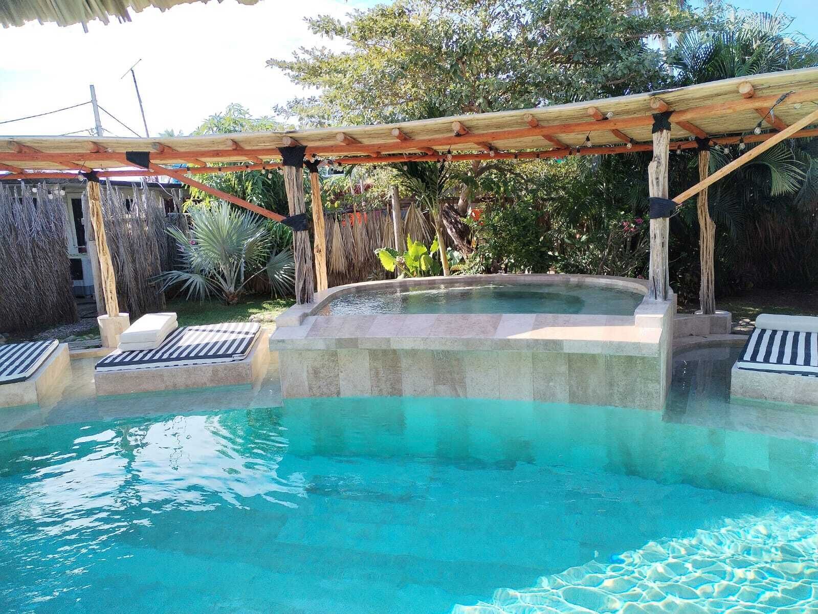 A swimming pool with a wooden pergola overhead, surrounded by lush greenery.