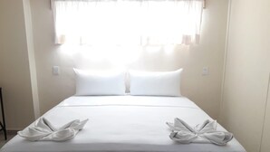 Una cama bien tendida con sábanas y almohadas blancas, una ventana con cortinas traslúcidas y una mesita de noche sencilla.