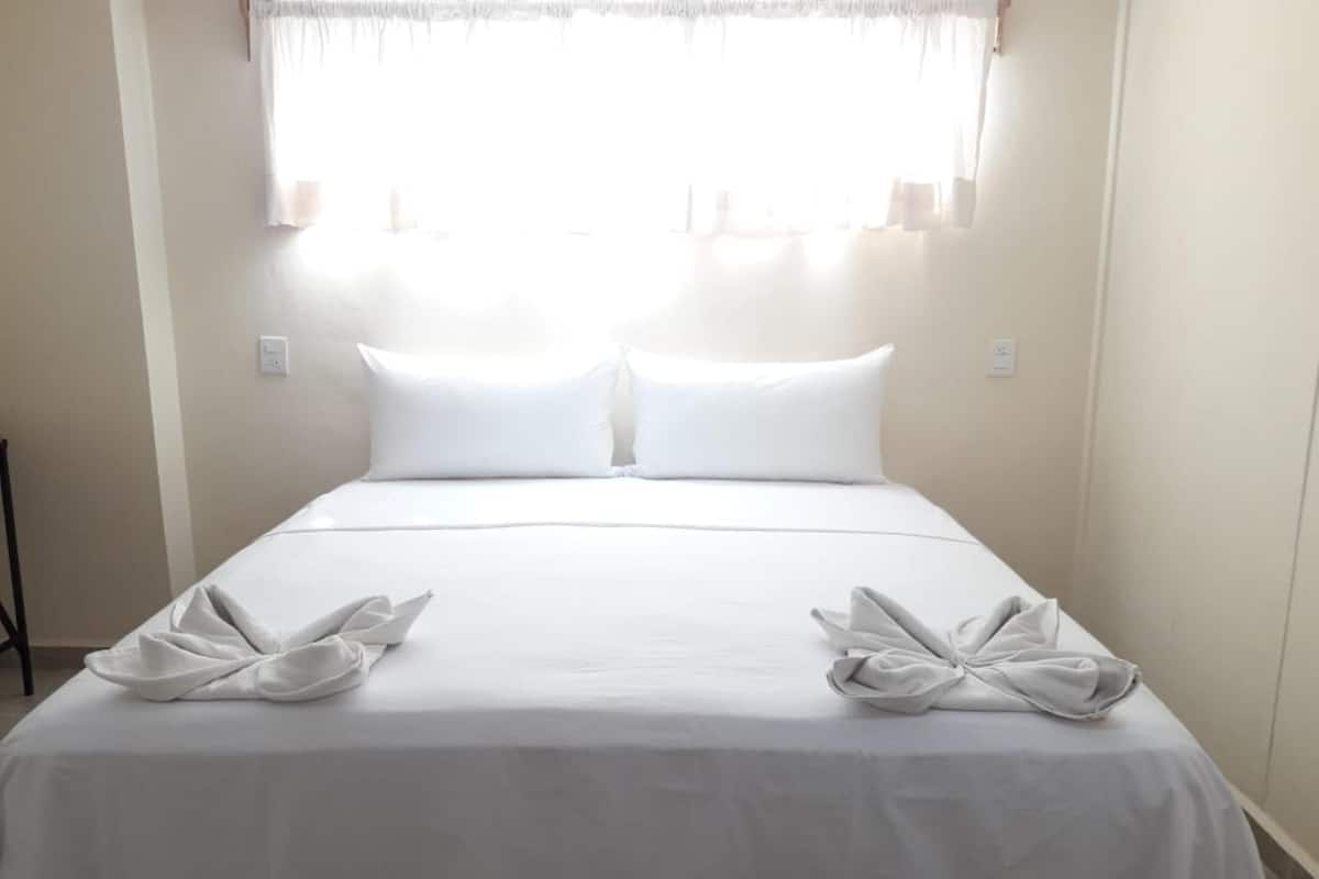 Una cama bien tendida con sábanas y almohadas blancas, una ventana con cortinas traslúcidas y una mesita de noche sencilla.