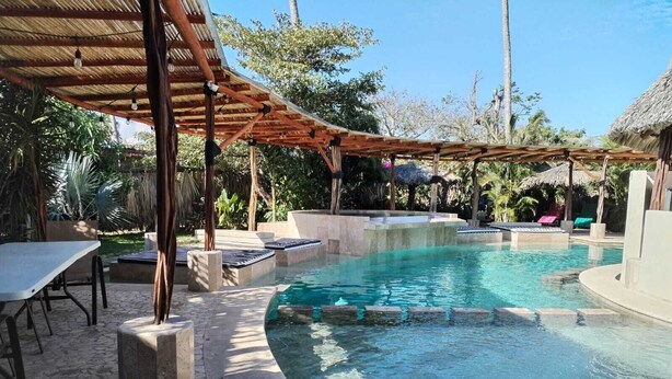 Zona de piscina con borde curvo, sillas de descanso y un área de estar cubierta.