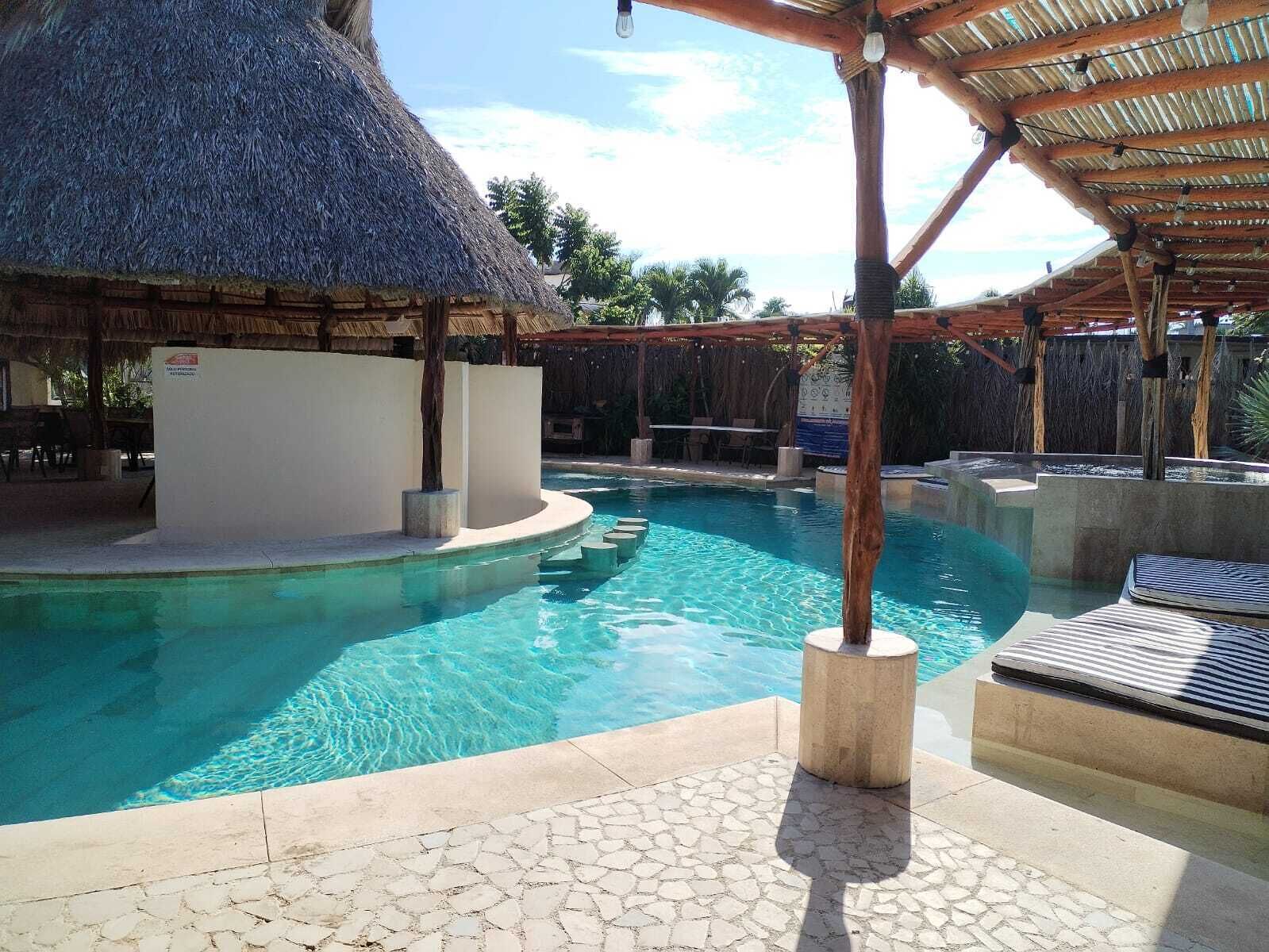 Ein Pool mit strohgedeckter Struktur, einer Fliesen-Terrasse und einer Liegebereich mit Sonnenliegen.