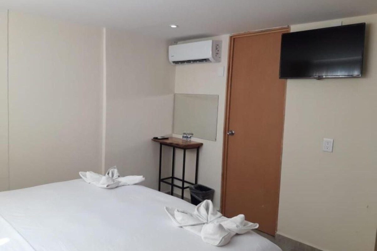 Habitación de hotel con cama, televisor montado en la pared, equipo de aire acondicionado, una mesita con espejo y una puerta.