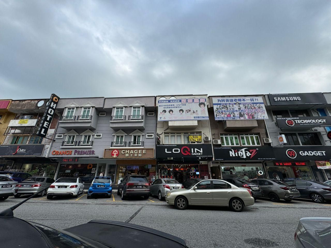 A street view with multiple commercial buildings, including a hotel, a store named 'Orange Premier,' and a 'Chaggee' store.