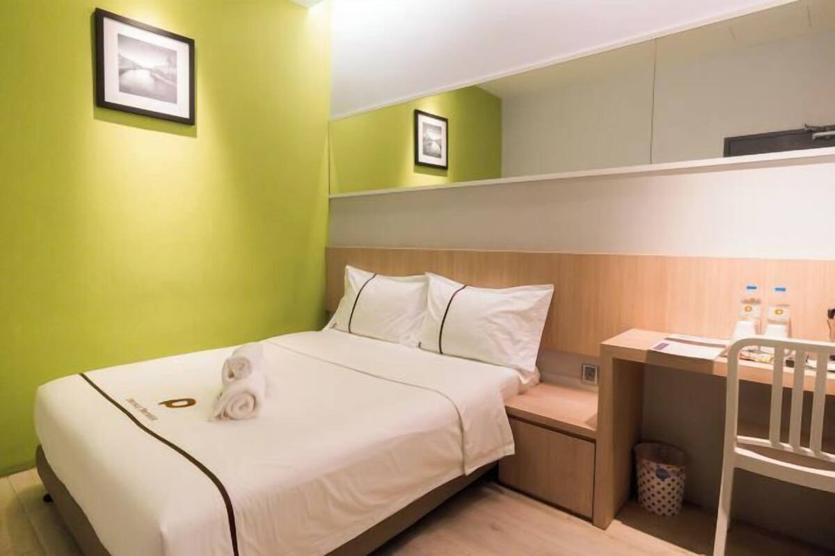 A hotel room with a bed, a desk, a chair, and a trash bin.