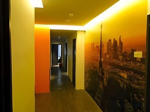 Un couloir orné d’une fresque représentant un paysage urbain sur un de ses murs, avec un luminaire au plafond et une porte au fond.