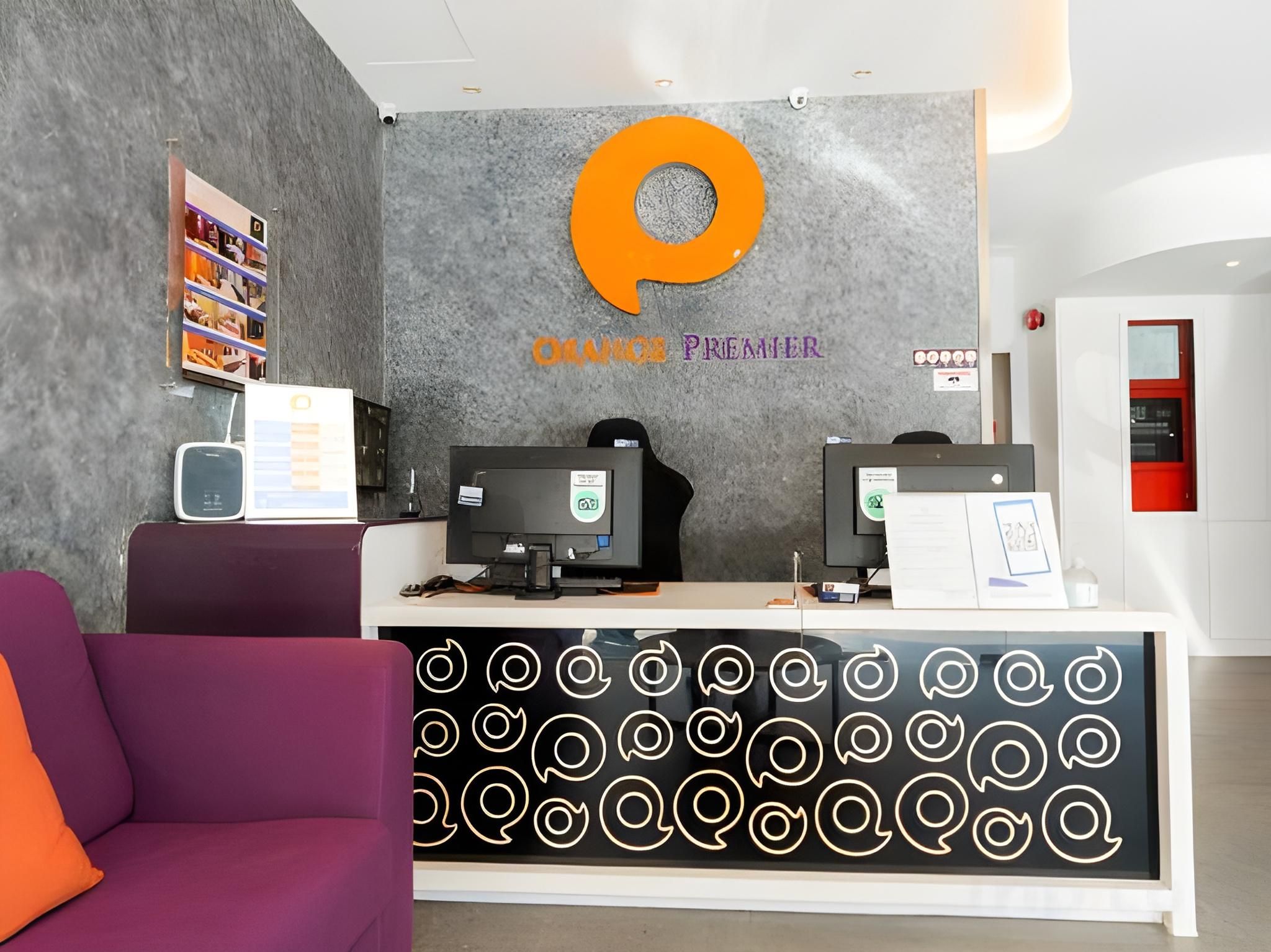 A reception area with a modern design, featuring a large orange logo, two computer monitors, and a seating area with a purple couch.