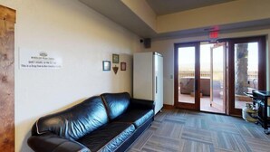 A room with a black leather sofa, a refrigerator, a kitchen area with a stove, and a sliding glass door leading to an outdoor patio. - Large Groups | WiFi | Wheeler Peak Lodge South (Angel Fire)