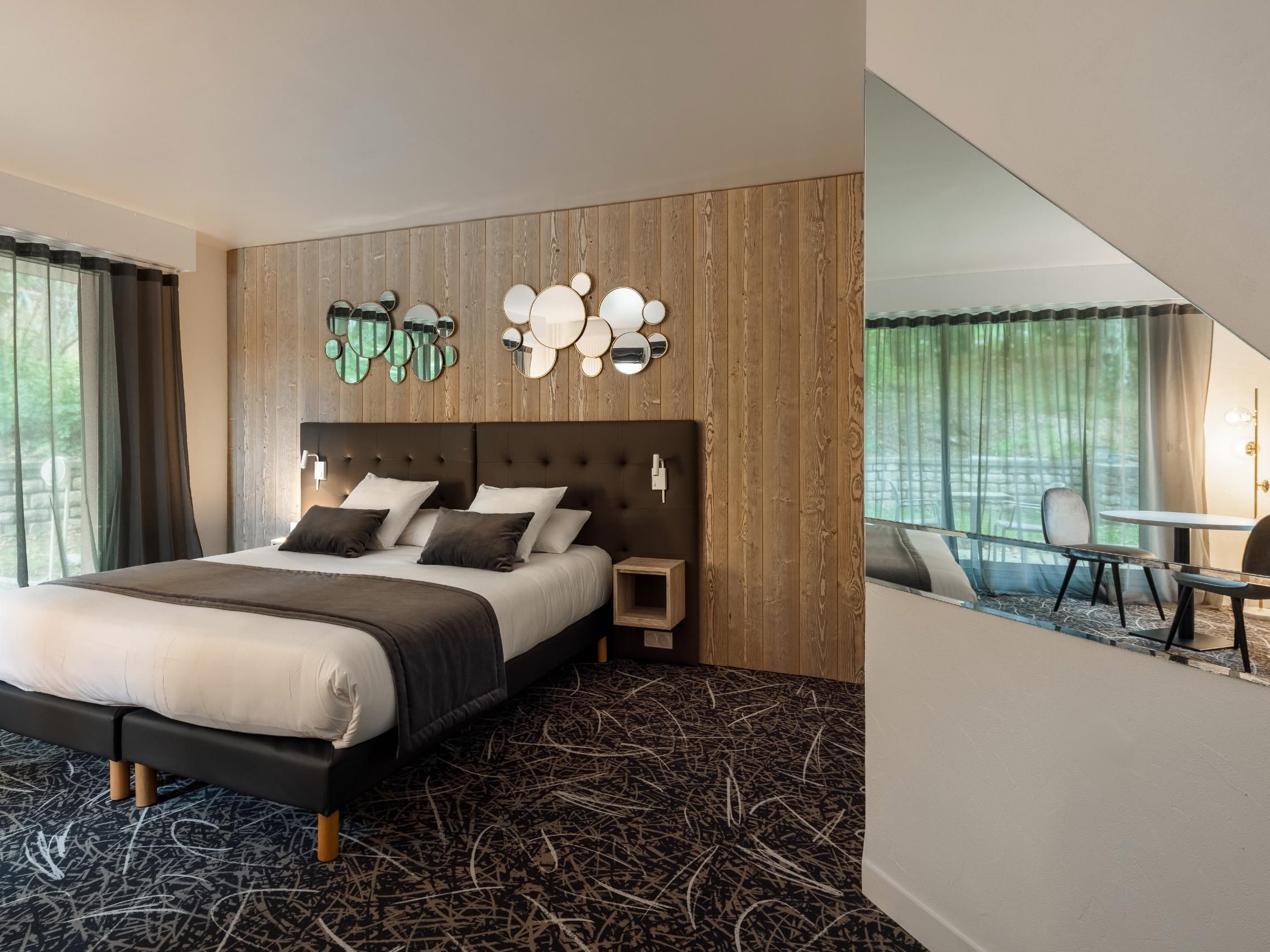 Una habitación de hotel moderna con una cama grande, un cabecero de madera, un espejo montado en la pared y una pequeña mesa con sillas.