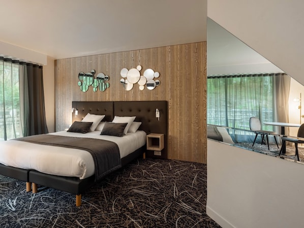 A modern hotel room with a large bed, a wooden headboard, a wall-mounted mirror, and a small table with chairs.
