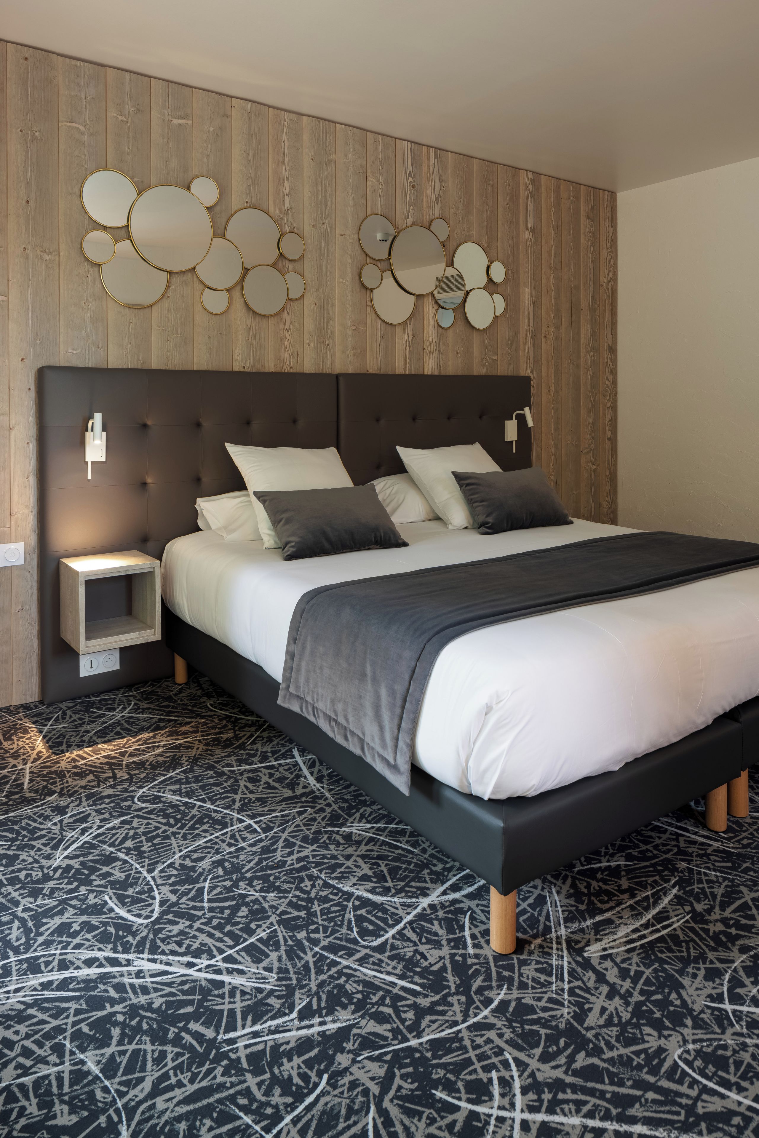 A modern bedroom with a tufted headboard, a bed with white and dark gray bedding, a bedside table, and a wall-mounted mirror.