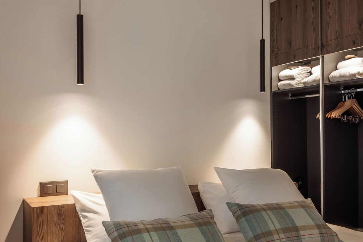 A modern bedroom with a wooden headboard, a large bed with plaid bedding, a nightstand with a lamp, and a wardrobe with hanging clothes.