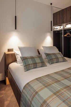 A modern bedroom with a wooden headboard, a large bed with plaid bedding, a nightstand with a lamp, and a wardrobe with hanging clothes. - Logis Hotel Spa Parenthese (Chille)