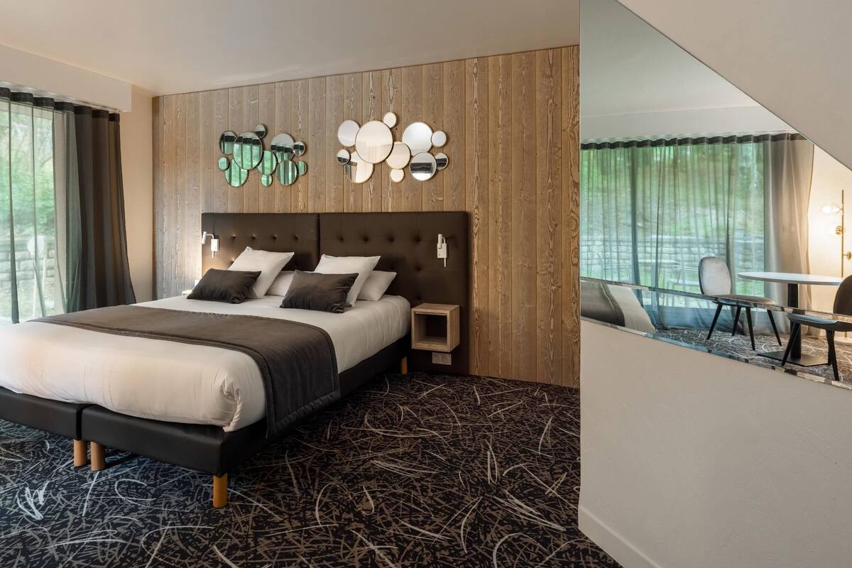 Una habitación de hotel moderna con una cama grande, un cabecero de madera, un espejo montado en la pared y una pequeña mesa con sillas.