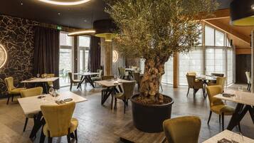 Interior de restaurante con mesas redondas, sillas, una gran oliva en maceta e iluminación moderna.