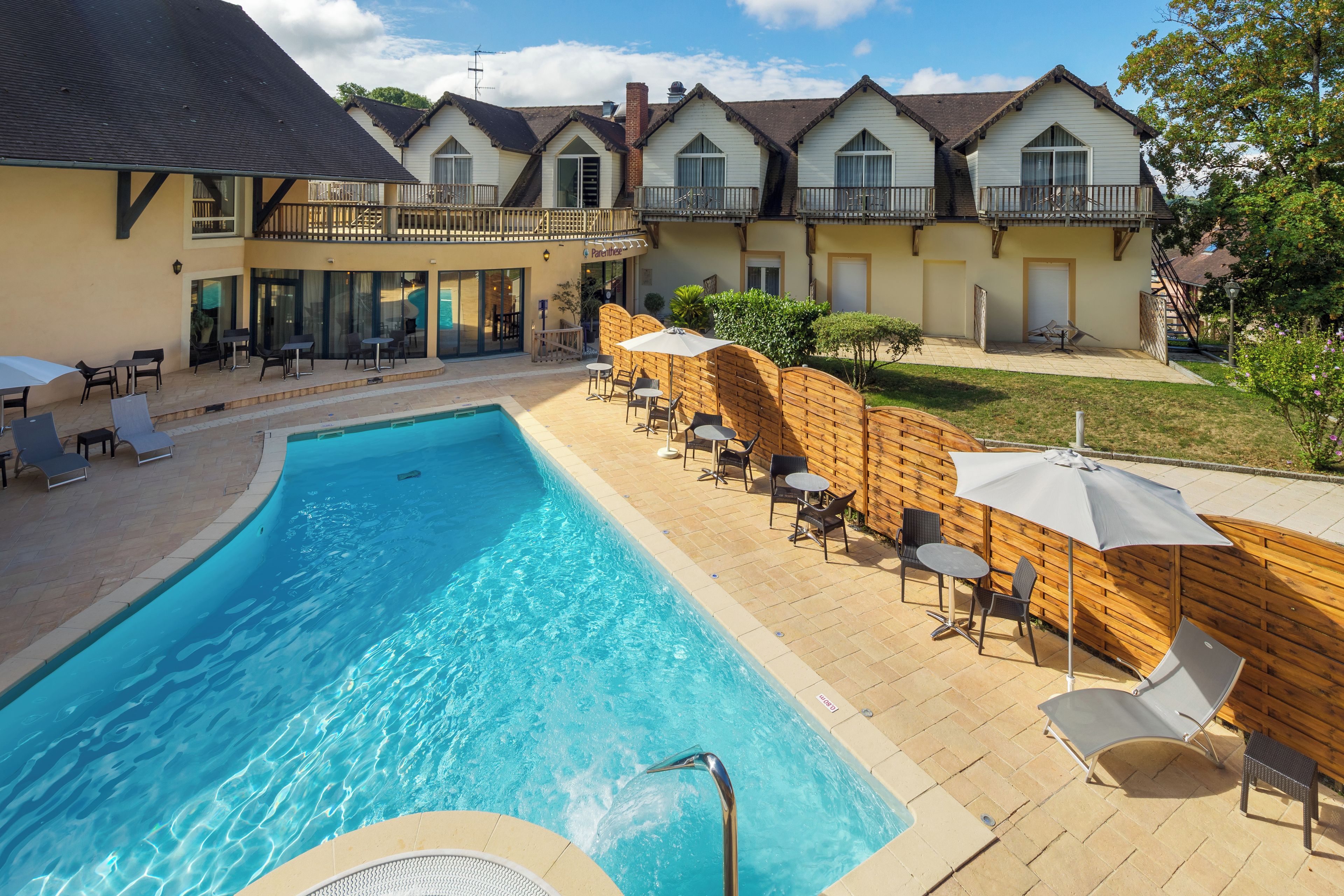 Una piscina con sillas de descanso y sombrillas, rodeada de cercas de madera y un edificio con balcones.