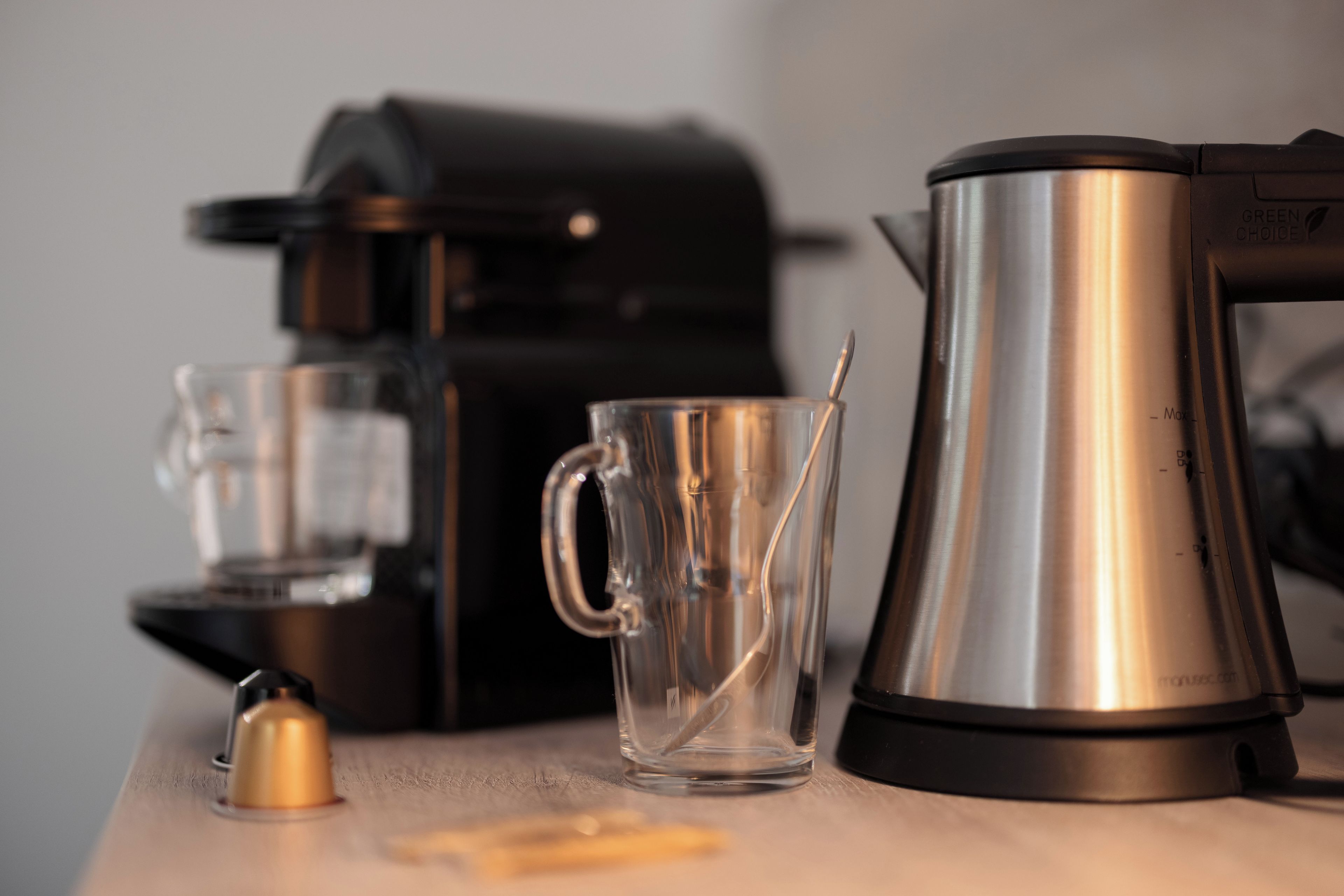 A coffee maker, a glass mug, and a spoon on a wooden table.