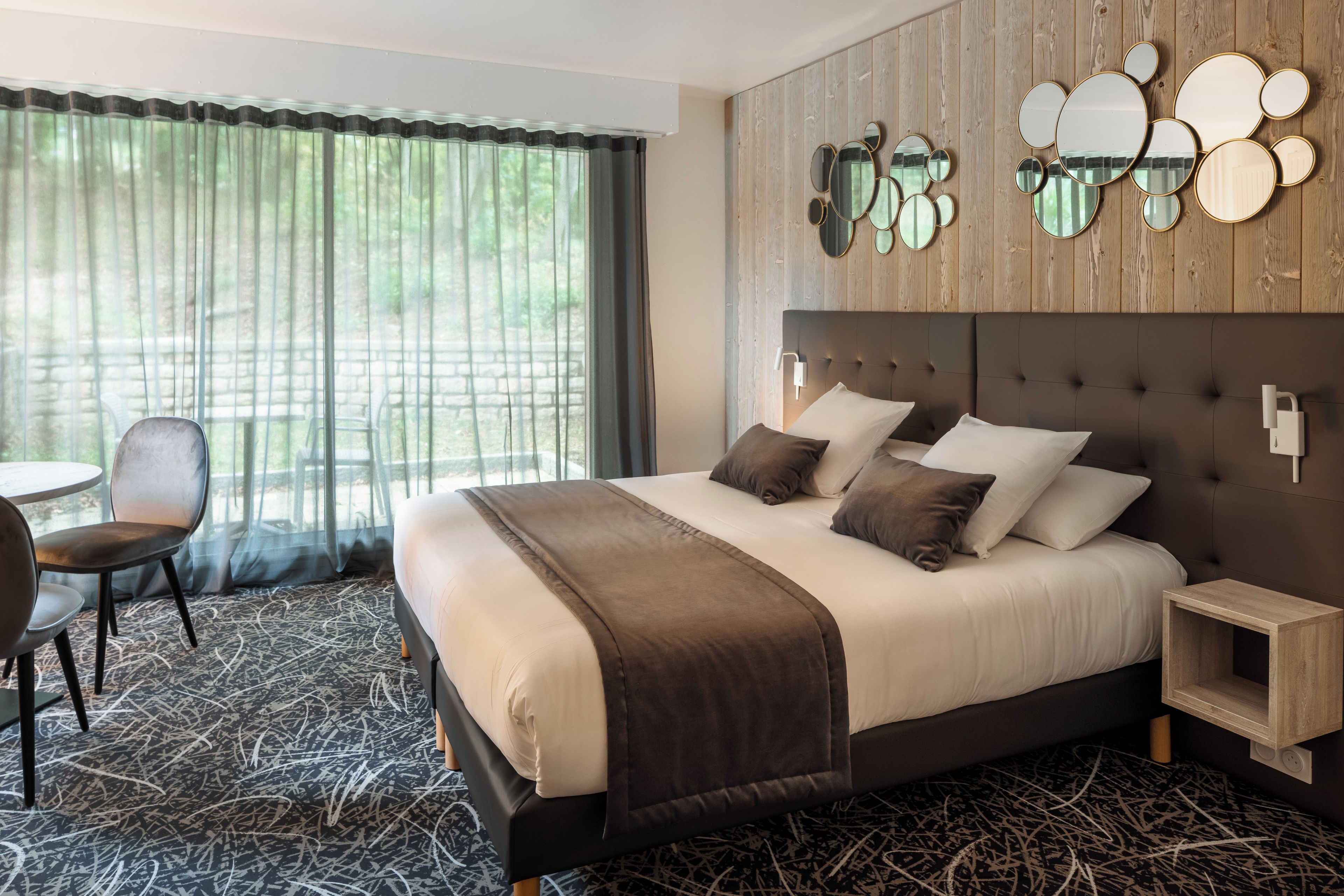 A hotel room with a large bed, a chair, a small table, and a view of greenery through the window.