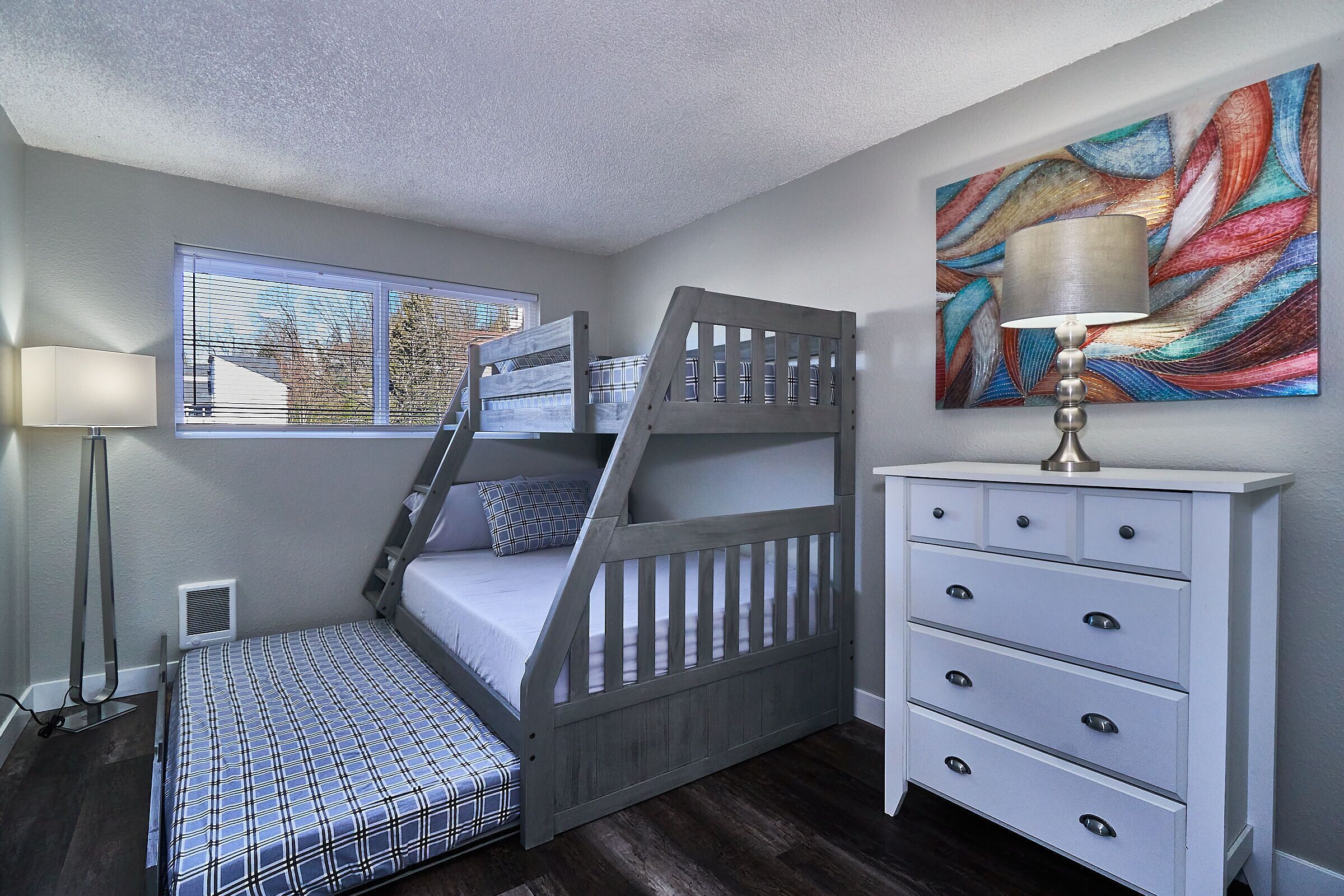 A bedroom with a bunk bed, a bedside table with drawers, a lamp, and a window with blinds.
