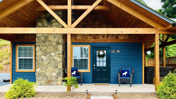 Une cabane avec des fondations en pierre, des poutres en bois, une porte bleu foncé et deux fauteuils en osier dotés de coussins à motifs d’élans.