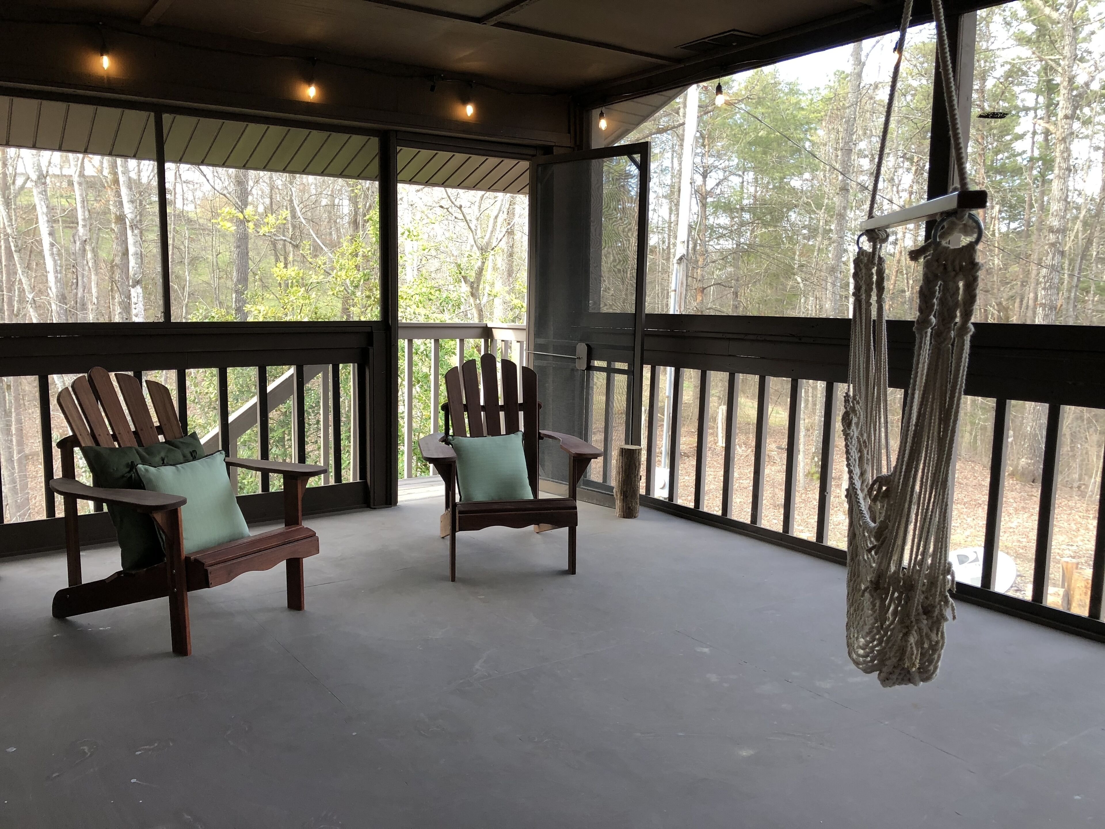 A covered porch with two Adirondack chairs, a hammock, and a view of a wooded area.
