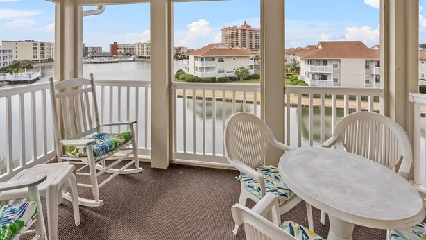 A covered balcony with white chairs and a table, overlooking a body of water with buildings in the background. - Cozy villas near the heart of North Myrtle Beach (North Myrtle Beach)