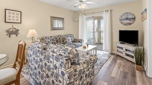 A living room with a patterned sofa, a ceiling fan, a television, and a dining area with a table and chairs. - Cozy villas near the heart of North Myrtle Beach (North Myrtle Beach)