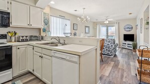 A modern kitchen with white cabinets, a dishwasher, a stove, and a microwave. There is a dining area with a patterned table and chairs, and a living room with a TV and a clock on the wall. - Cozy villas near the heart of North Myrtle Beach (North Myrtle Beach)