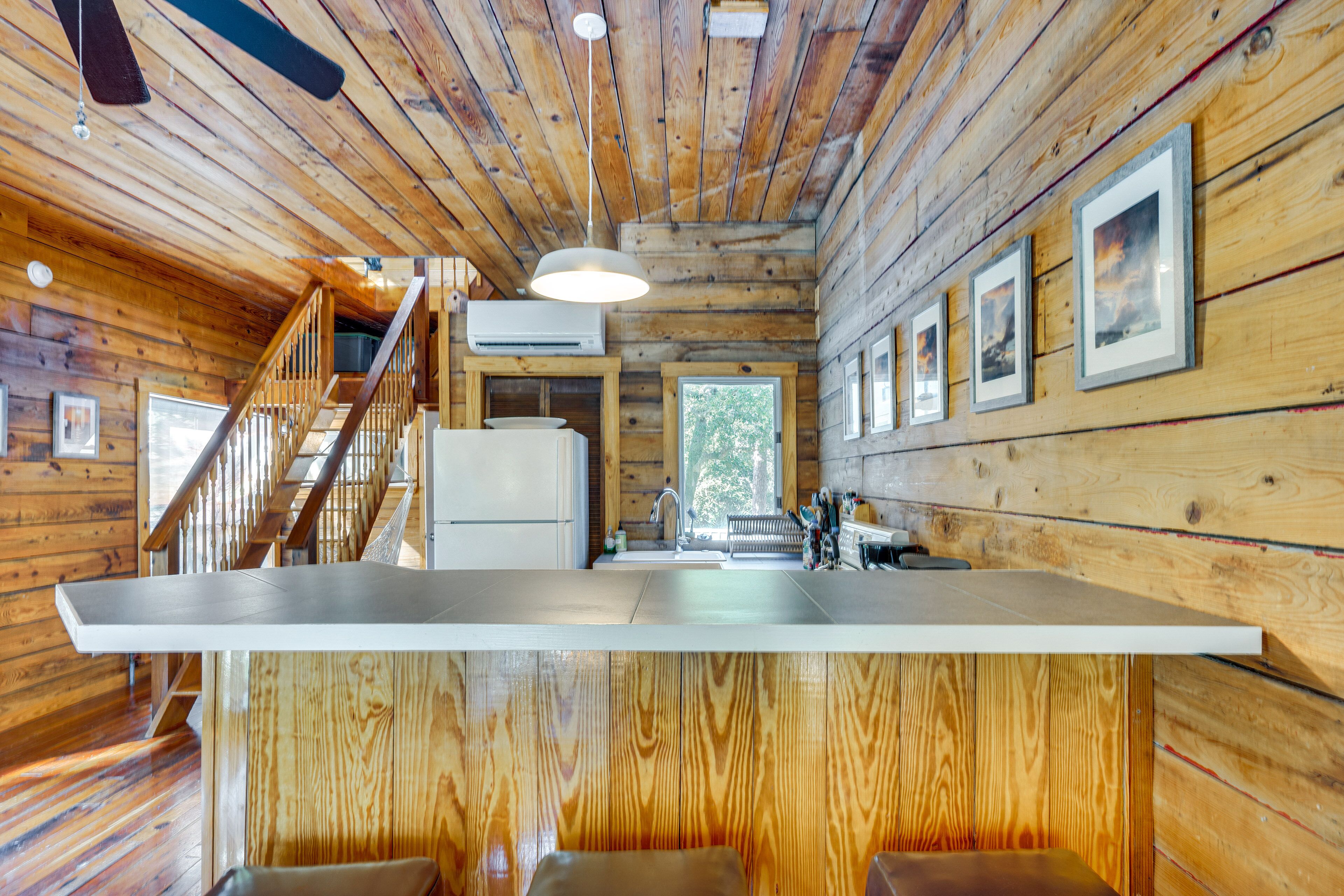 A wooden cabin interior with a kitchen area, wooden walls, a staircase, and framed pictures on the wall.