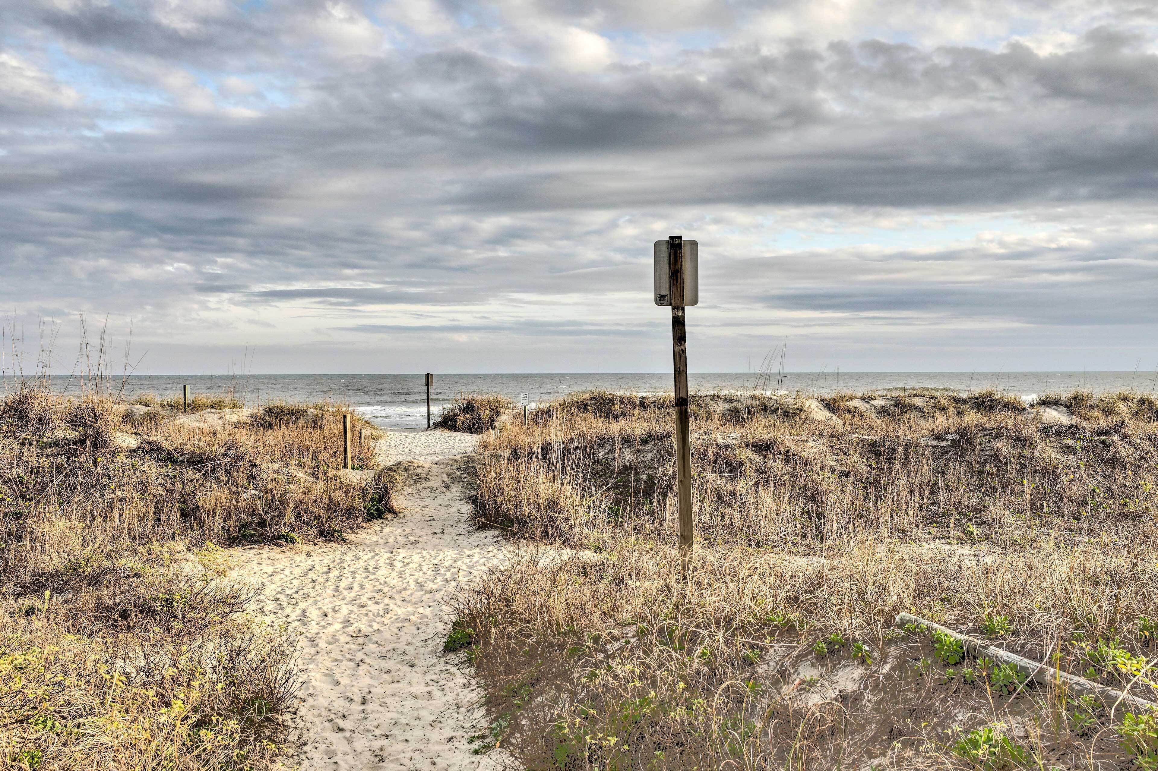A sandy beach path leading to the sea, with a weathered signpost in the distance.
