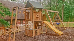 A wooden play structure with a slide and climbing elements, surrounded by a fence and mulch.