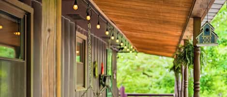 A wooden porch with a swing chair, hanging lights, and a birdhouse.