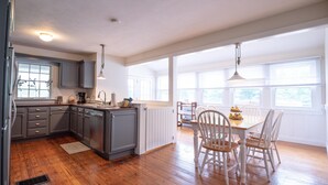 Une cuisine avec des meubles de couleur grise, un plancher en bois et un coin repas avec des chaises et une table blanches.