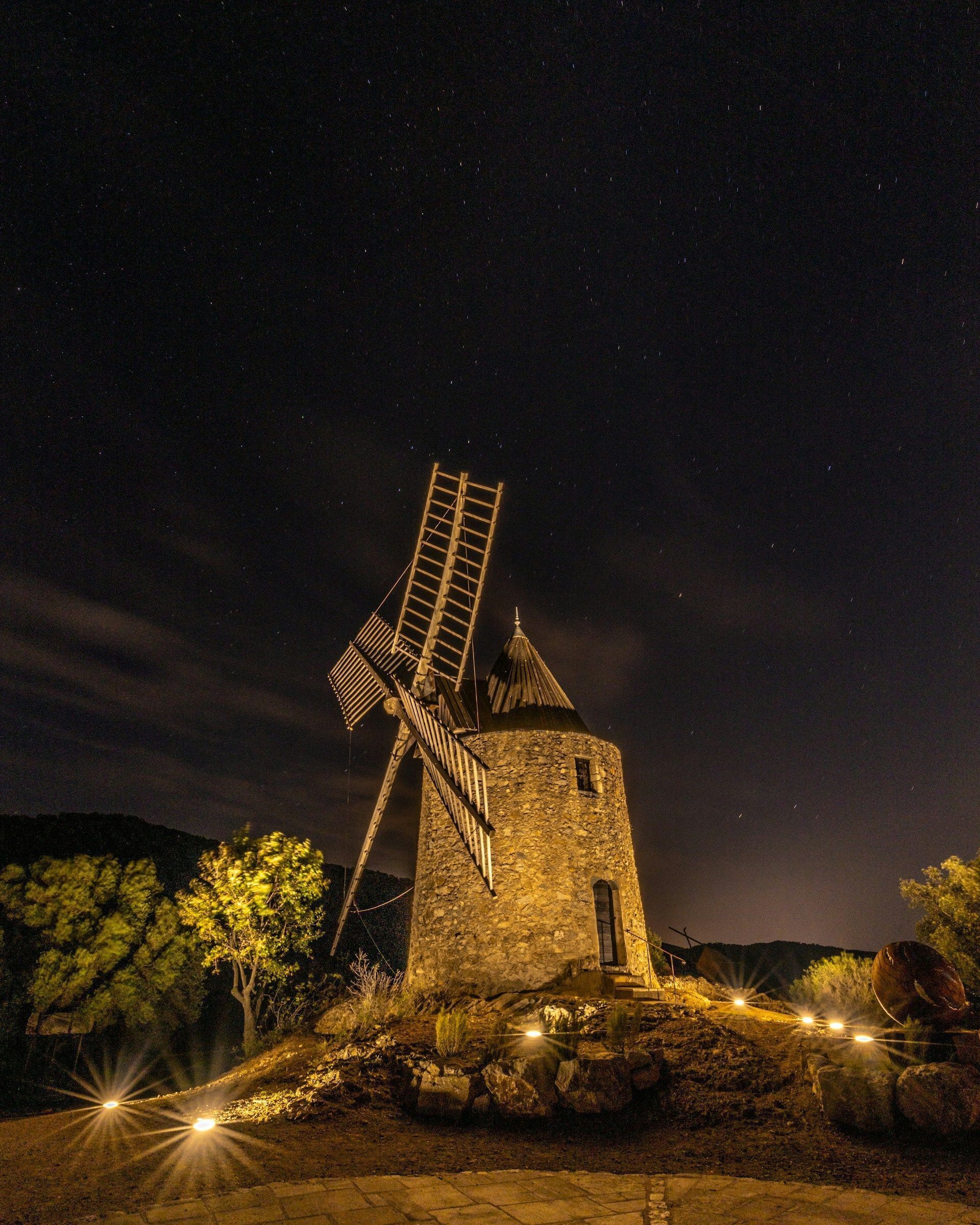 Een stenen windmolen in de nacht met een ladder, omringd door rotsen en bomen.