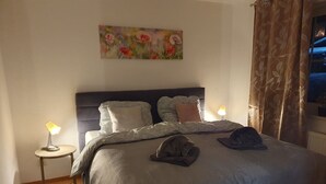 A bedroom with a bed, two bedside lamps, a painting on the wall, and a window with curtains.