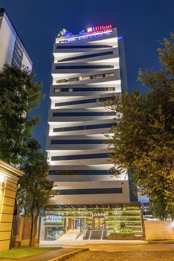 Un hotel moderno con un llamativo letrero de Hilton Garden Inn, varios pisos y una entrada bien iluminada con jardín paisajístico.
