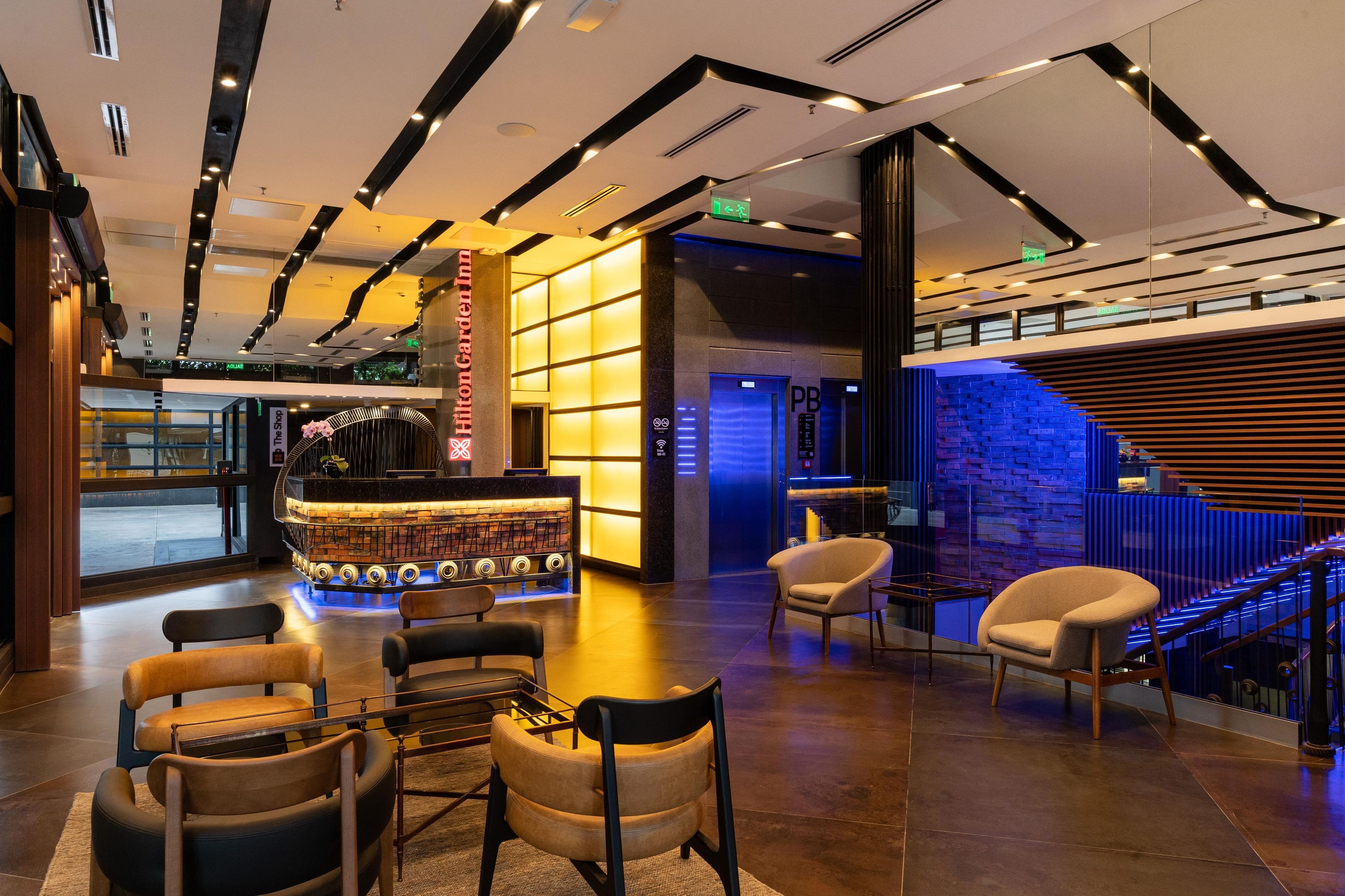 A modern hotel lobby with a reception desk, seating area, and a staircase with a blue-lit wall.