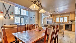 A spacious dining area with a wooden table and chairs, a large window, and a kitchen with wooden cabinets and a refrigerator. - Hot Tub Relaxation: Breck Gem 3 Mi to Skiing! (Breckenridge)
